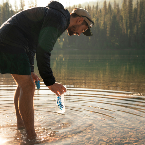 A person refilling a water bottle from a lake while hiking